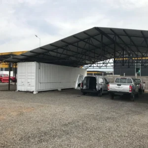 Contenedor blanco bajo galpón con estructura metálica y vehículos estacionados